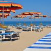 Spiaggia vuota con lettini blu e ombrelloni arancioni, passerella bianca e blu verso il mare.