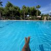 Gambe in una piscina turchese con palme e cielo azzurro sullo sfondo.