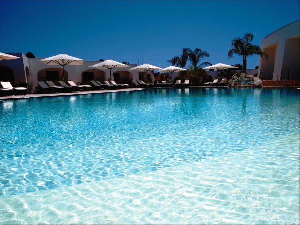 Piscina all'aperto con lettini e ombrelloni bianchi, cielo limpido e palme sullo sfondo.